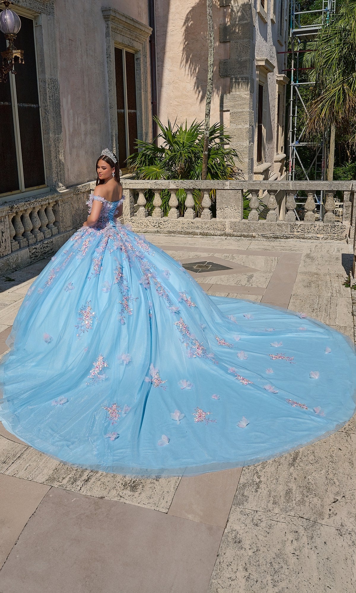 A woman in the Quinceanera Dress 54228 By Amarra, an off-the-shoulder blue ball gown with pink 3D floral appliqué and a tiara, stands on a stone terrace outside a historic building.